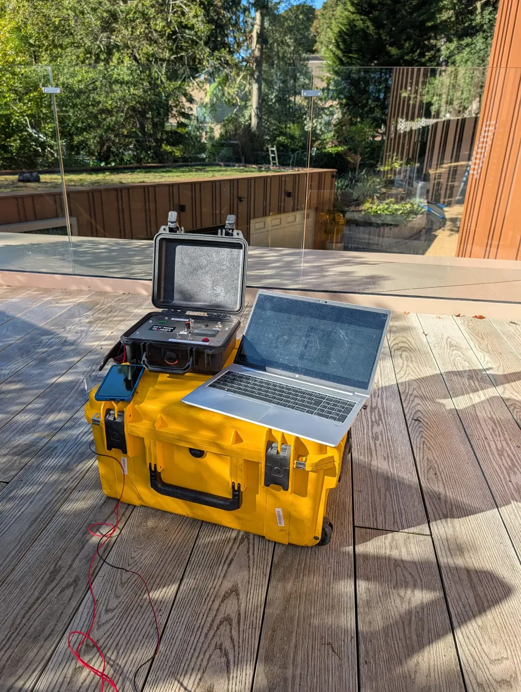 Buried leak location survey set-up on a roof terrace beside a green roof: field generator case, laptop and phone on a transport case, connecting lead run across the decking