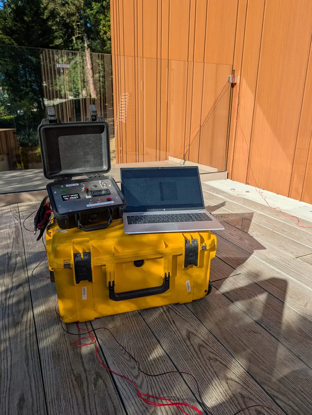 Buried leak location survey equipment set up on a roof terrace: field generator in a protective case with a laptop on top, and the red connecting lead run to the building